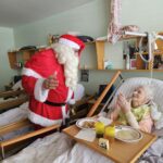 Božiček stoji ob postelji stanovalke, ki z nasmehom in sklenjenimi dlanmi sedi pri obroku.