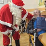 Božiček stoji ob stanovalcu z hojico in mu izroča darilo v skupnem prostoru doma.