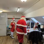 Božiček in Božička obiskujeta zaposlene v pisarni doma; zaposleni sedijo za mizo in se smehljajo.