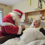 Božiček se skloni k stanovalki v postelji in ji izroča darilo v sobi doma za starejše.