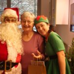 Stanovalka pozira z Božičkom in palčico ob božično okrašenem drevesu.