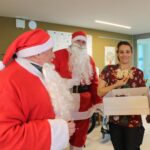 Dva Božička stojita ob zaposleni, ki drži škatlo z darili, in skupaj se nasmihajo v notranjem prostoru doma.