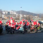 Skupina motoristov, oblečenih v Božičke, stoji z motorji na parkirišču pred domom; v ozadju so zasnežena polja in vas.