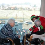 Dve stanovalki sedita ob velikem oknu, med njima je palčica v božičnem kostumu, ki se z njima pogovarja; spodaj na parkirišču so motorji Božičkov.