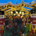 Skupina stanovalcev pozira v božični vasi, okrašeni z lučkami, oreščkarji in praznično dekoracijo.