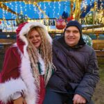 Stanovalec sedi na klopi v božični vasi poleg animatorke, obkrožen s praznično razsvetljavo in okraski.