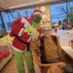 Oseba, preoblečena v Grincha v rdeče-zeleni kostumski opravi, stoji ob starejši stanovalki in jo drži za roko v skupnem prostoru doma.