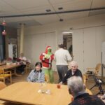 Grinch v božični preobleki stoji med mizami v jedilnici doma za starejše, okoli njega pa sedijo stanovalci in opazujejo dogajanje.