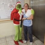 Grinch, oblečen v božični kostum, pozira skupaj z zaposleno v delovni uniformi ob oglasni deski v notranjem prostoru doma.