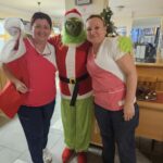 Grinch v božični preobleki stoji med dvema zaposlenima v domu za starejše in se z njima nasmejano fotografira v hodniku.