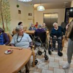 Skupina starejših stanovalcev sedi za mizami v skupnem prostoru in spremlja glasbeni nastop, v ospredju so pripomočki za hojo.