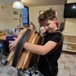 Otrok od blizu igra harmoniko, v ozadju starejši stanovalci ploskajo in z zanimanjem spremljajo nastop.