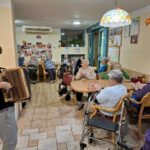 Otrok z harmoniko nastopa pred zbranimi stanovalci doma, ki sedijo v polkrogu in ploskajo v skupnem prostoru.