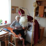 Oseba, oblečena v kostum sv. Miklavža, obiskuje stanovalca v postelji v sobi doma za starejše.