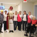 Skupinska fotografija sv. Miklavža z zaposlenimi in stanovalcem v invalidskem vozičku na hodniku doma za starejše.