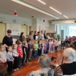 Skupina otrok in vzgojiteljic stoji v vrsti ter nastopa pred starejšimi stanovalci, ki sedijo v jedilnici doma za starejše.