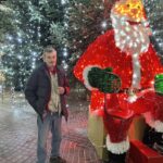 Moški stoji ob veliki osvetljeni figuri Božička na večerno okrašenem trgu z božičnimi lučkami.