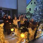 Skupina starejših stoji na praznično okrašenem saneh z lučkami in božičnim okrasjem na prostem.