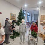 Skupina starejših oseb skupaj postavlja umetno božično drevo; nekateri držijo veje, drugi jih pritrdijo ali prinašajo nove kose.