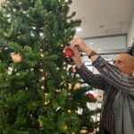 Starejši moški obeša rdečo bleščečo kroglico na že deloma okrašeno božično drevo z lučkami.