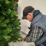 Starejši moški v kapi namešča svetlobni kabel na spodnji del božičnega drevesa, medtem ko drugi stoji za drevesom in pomaga pri okrasitvi.