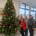 Skupina stanovalcev in stanovalka z animatorko ponosno pozirajo ob popolnoma okrašenem božičnem drevesu z rdečimi in zlati okraski.