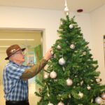 Starejši moški z rjavim klobukom obeša okraske na božično drevo na hodniku doma za starejše.