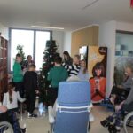 Otroci okrašujejo božično smrečico v skupnem prostoru doma, okoli njih sedijo stanovalci v invalidskih vozičkih.