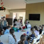 Otroci in stanovalci doma sedijo za mizami v jedilnici, kjer jim odrasla oseba ponuja krožnik s pecivom.