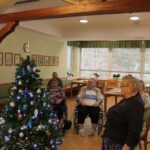 Skupina stanovalcev doma za starejše stoji in sedi ob okrašeni božični smrečici v jedilnici.