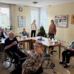 Skupina starejših stanovalcev sedi za mizami v dnevnem prostoru doma; nekateri uporabljajo invalidske vozičke ali hodulje, v ozadju stojita dve mlajši obiskovalki ob okrašeni božični smrečici.