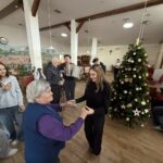 Mlada obiskovalka pleše s starejšo stanovalko v skupnem prostoru doma; v ozadju so drugi stanovalci, mladi glasbeniki z harmoniko ter okrašena božična smreka.