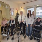 Skupinska fotografija starejših stanovalk z uporabo hodulj in mlajših obiskovalk, zbranih ob okrašeni božični smrečici v notranjem prostoru doma.