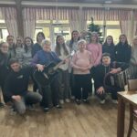 Velika skupina mladih in starejših udeležencev stoji skupaj v skupnem prostoru doma; v ospredju starejša stanovalka z glasbilom, ob njej mladi z harmoniko, v ozadju praznično okrašena smrečica.