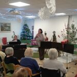 Operna pevka nastopa pred stanovalci doma, ki sedijo v dvorani, okrašeni z božično smreko in prazničnimi lučkami.