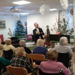 Operni pevec govori ali poje pred občinstvom stanovalcev, v ospredju so invalidski vozički, v ozadju praznična dekoracija.