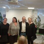 Skupinska fotografija štirih nastopajočih pred božično okrašeno dvorano z glasbenimi stojali.