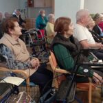 Stanovalci doma sedijo in poslušajo glasbeni nastop, nekateri uporabljajo pripomočke za hojo.