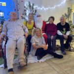 Skupina stanovalcev in zaposlenih pozira ob božični dekoraciji; med njimi je oseba v kostumu Božička, okoli pa so okrašena drevesca in praznične lučke.