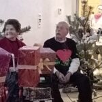 Dva stanovalca sedita za mizo z zavitimi darili in božično dekoracijo; v ozadju je okrašeno notranje drevo.