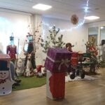 Praznično okrašen prostor z velikimi darili, snežakom in božičnimi figurami; prisotni so otroci in odrasli.