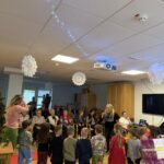 Otroci stojijo pred starejšimi stanovalci v dnevnem prostoru, okrašenem za praznike, medtem ko vzgojiteljica vodi nastop.