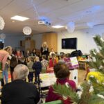 Otroci nastopajo v praznično okrašenem prostoru doma, starejši stanovalci sedijo v ozadju in spremljajo dogajanje.