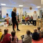 Otroci sedijo na blazinah in spremljajo praznično dogajanje, v ospredju so darila in praznični okraski, v ozadju stojijo zaposleni in stanovalci.