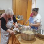 Stanovalka pri mizi melje orehe v ročnem mlinčku, ob njej stoji zapos­lena in pomaga pri pripravi nadeva.