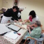 Starejše stanovalke in spremljevalka v predpasnikih skupaj razvaljajo testo in izrezujejo piškote za božične kekse za mizo v ustvarjalnem prostoru.