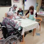 Skupina starejših stanovalcev sedi in stoji okoli mize ter sodeluje pri pripravi božičnih keksov z modelčki, valjarji in pekačem.