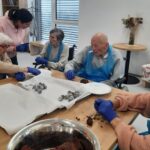 Starejši stanovalci in stanovalke sede za mizo oblikujejo čokoladne kroglice, ob pomoči zaposlene; na pladnju so pripravljene že oblikovane sladice.