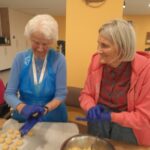 Dve stanovalki z modrimi predpasniki in rokavicami oblikujeta majhne piškote iz svetlega testa, nasmejani in v pogovoru.