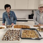 Dva stanovalca stojita za kuhinjskim pultom, pred njima so veliki pladnji pečenih in s čokolado okrašenih prigrizkov.